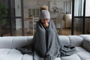 Cold Woman Sitting On Couch Wrapped In Blanket.